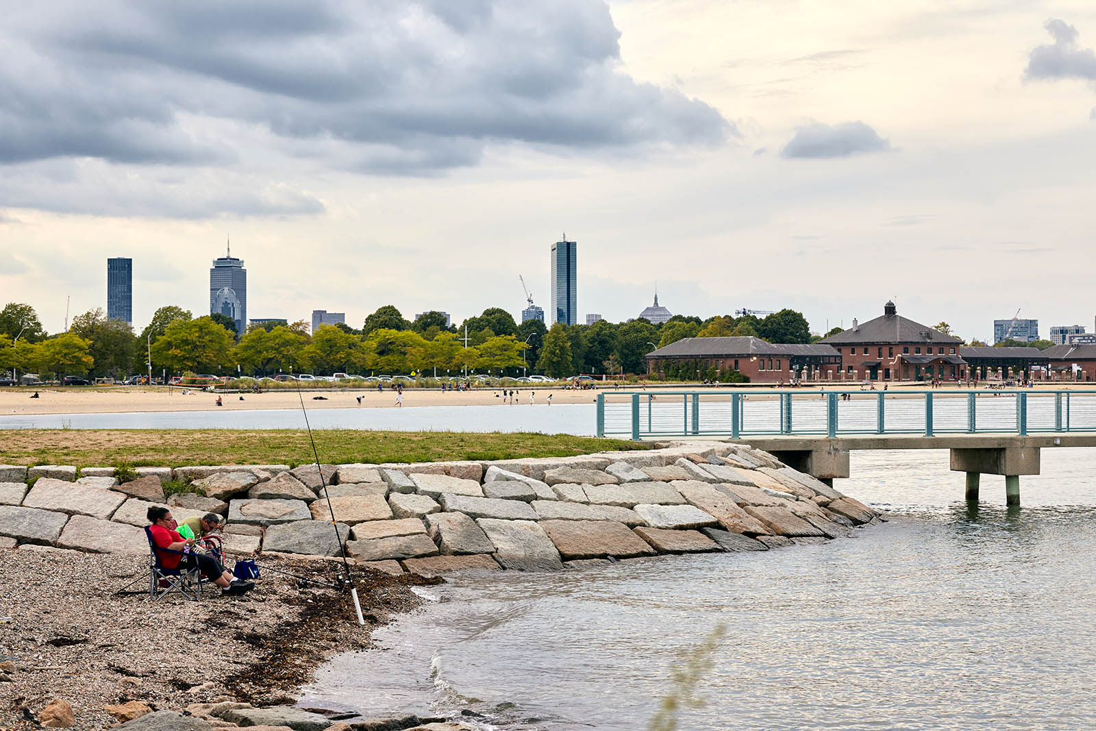 Photo of people fishing on the left side and a pier on the right side. Skyscarpers in the background