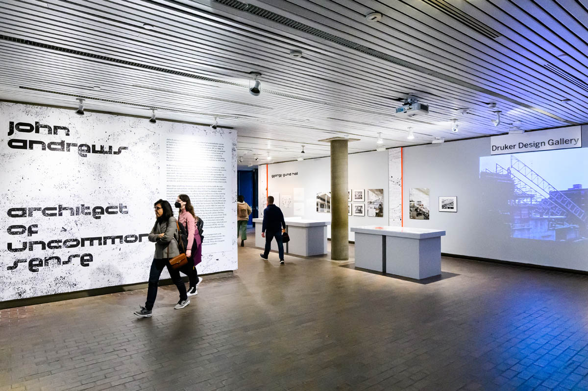 People walking around the exhibiiton title wall, display cases, and video projection.