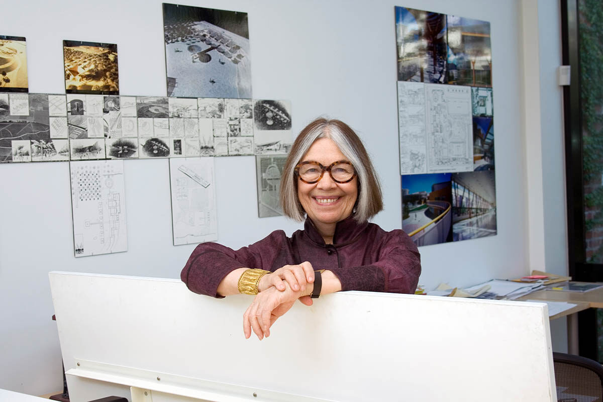 Adele Naude Santos sits in a studio with images on a wall behind her. She wears a dark purple shirt and glasses.