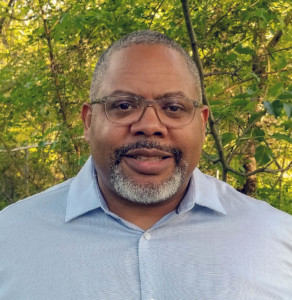 Headshot of Kofi Boone, who stands outside in front of leafy green trees.