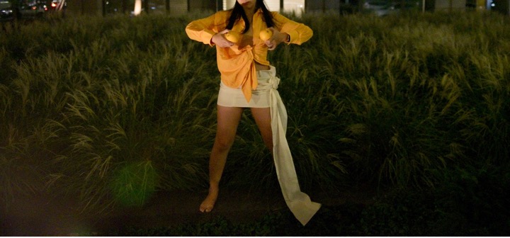 A woman wearing yellow and orange, stading on grass, holding an orange in each hand mimicing breasts.