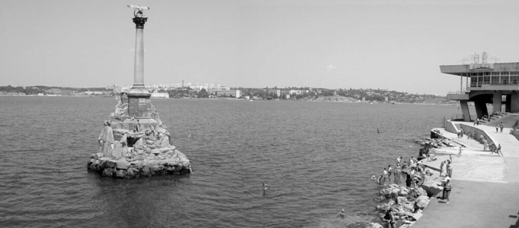 View of pillar monument in shallow bay; spectators and visitors on nearby shore.