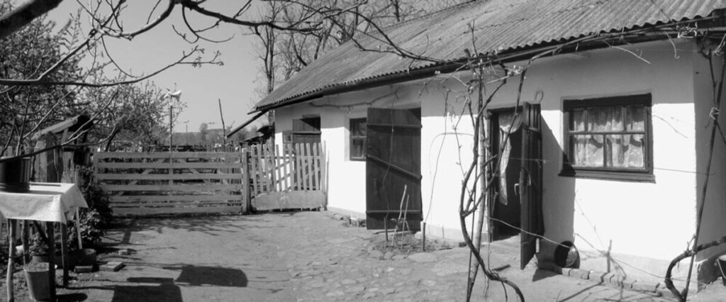 Photo of one-story white house with barren land in front, surrounded by wood fence.