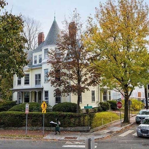 A white house with grey roof on the corner of the street, surrounded by trees.