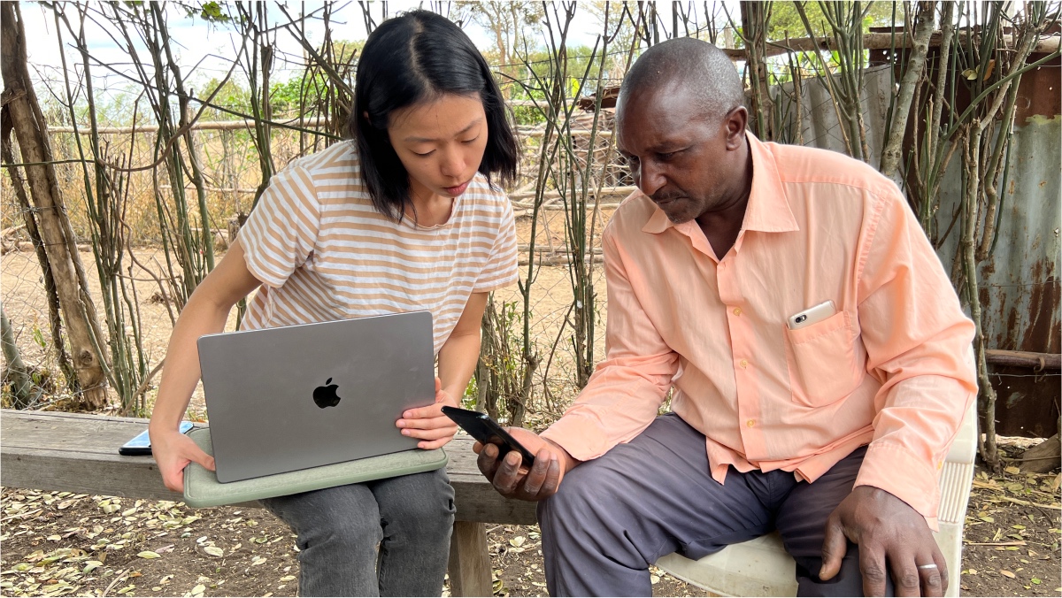 A photo of a Kenyan farmer and Caroline, a GSD student, using a smartphone and laptop while sitting on a bench.