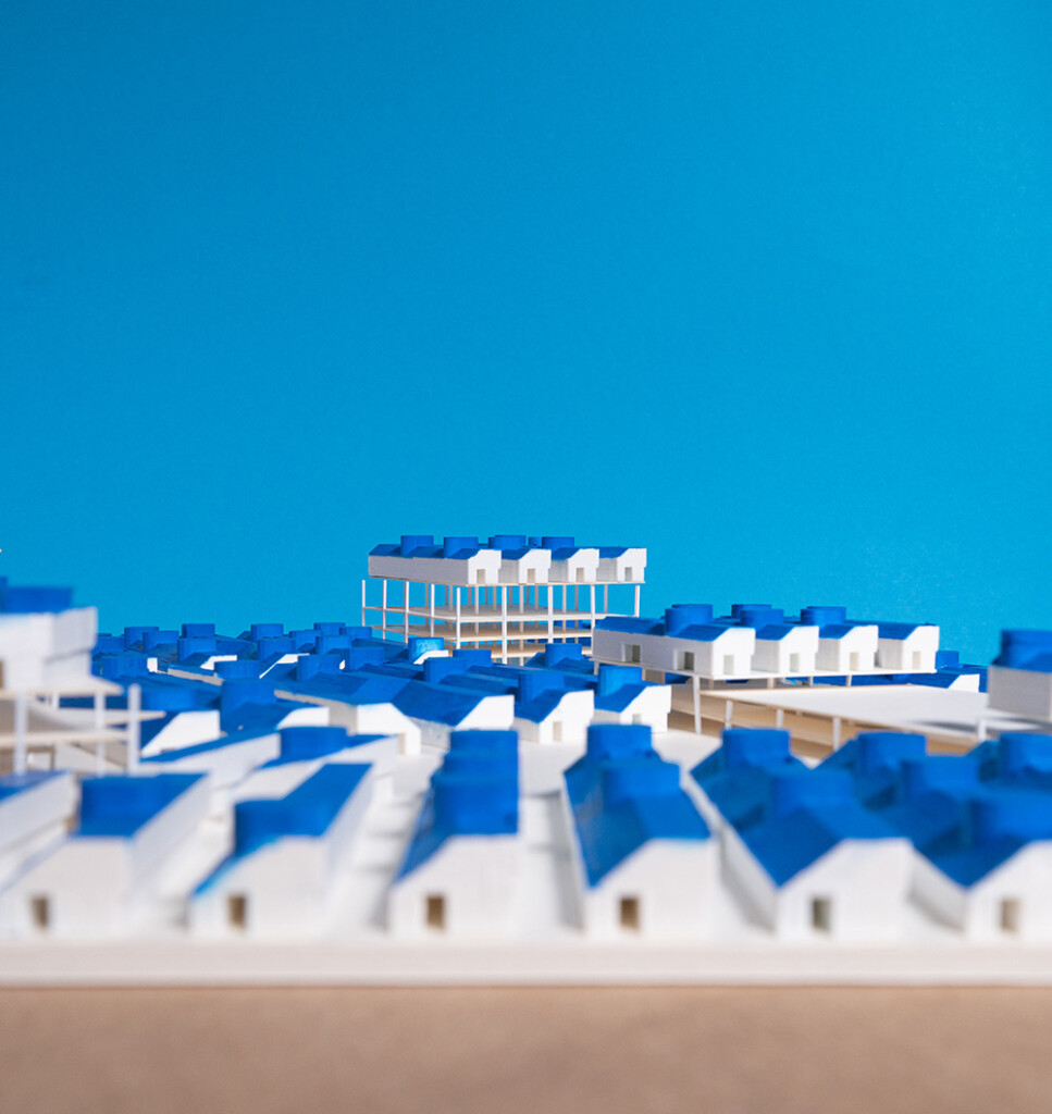 A depiction of an architectural model the features an array of small, uniform white structures with bright blue roofs arranged together on narrow alleys. Some structures are raided on plinth structures.