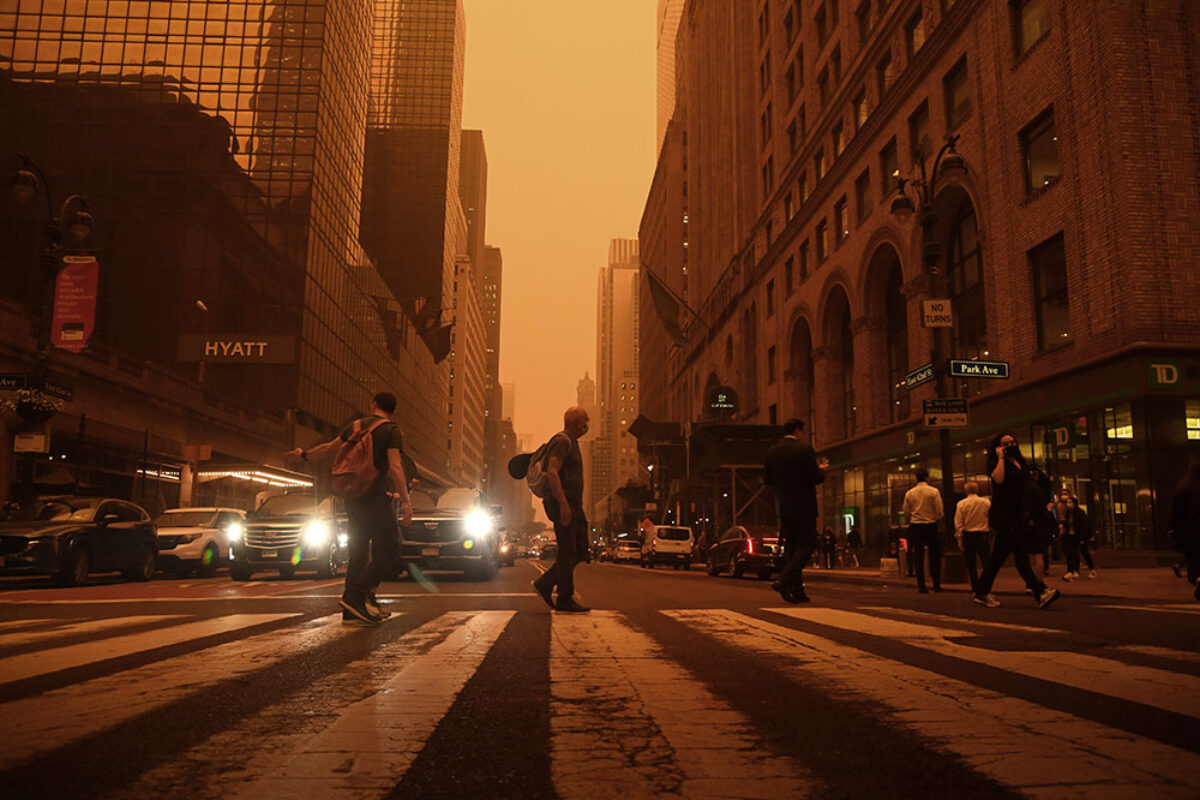 A photograph of a New York City street with pedestrians in a crosswalk and cars. The sky is orange grey and everything is tinted orange.