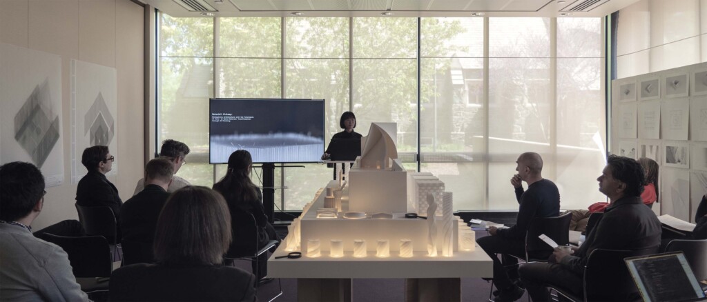 Sujie Park stands in front of a computer screen and several architectural models, presenting to a room full of people