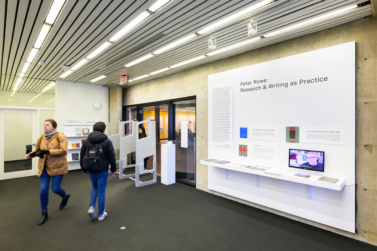 Two people walking past the introductory wall of books and their abstracts along with an interview of the author.