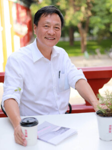 Portrait of Kongjian Yu, who wears a white shirt and sits at a table with a cup.