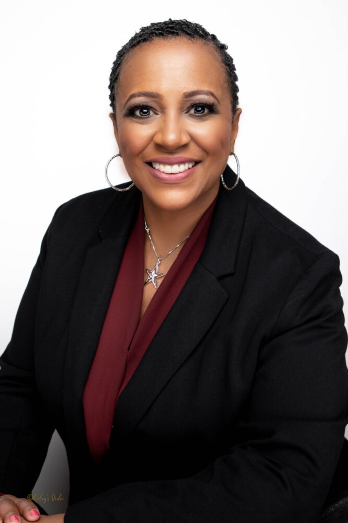 Portrait of Angela Brooks, who wears a black suit jacket, a maroon shirt, and hoop earrings.