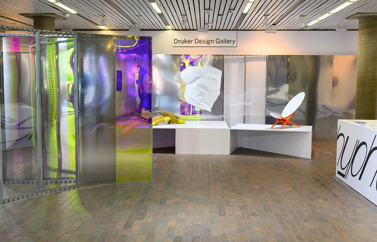 A view of Druker Design Gallery showing curved walls covered in reflective and colored plexiglass and tables displaying a variety of art objects.