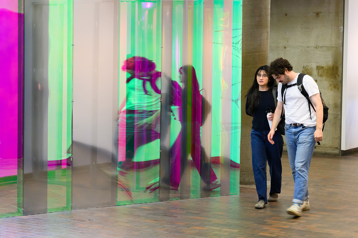 Two people walking in Druker Design Gallery with their images reflected in multiple colors on a curved plexiglass wall.