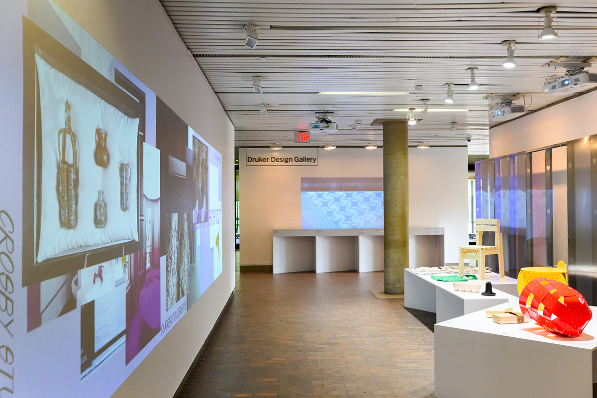 A view of Druker Design Gallery with projections on the walls and a variety of art objects on display tables.