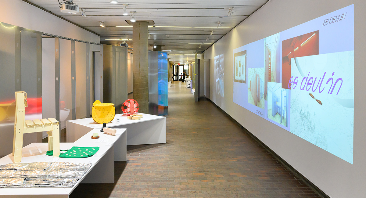 A view of Druker Design Gallery showing curved plexiglass walls, projections on flat walls, and a variety of art objects on display tables.