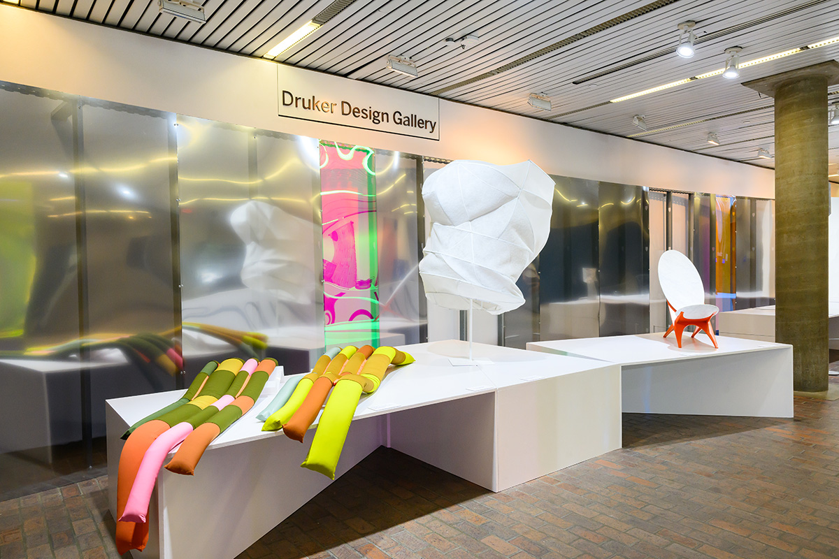 Tables display art objects in front of a reflective curved plexiglass wall inside of Druker Design Gallery.