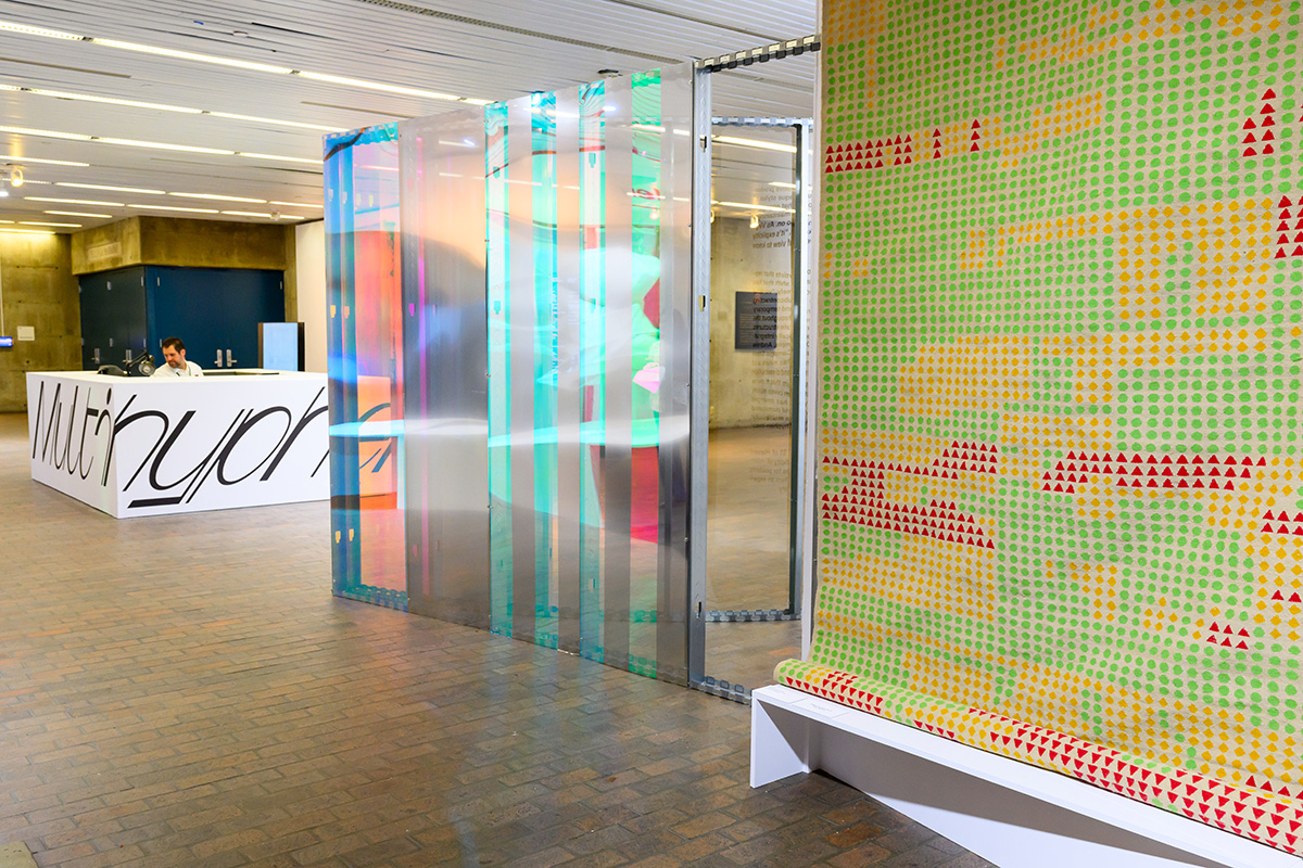 A large tapestry with a colorful geometric design displayed in front of a curved reflective plexiglass wall inside Druker Design Gallery.