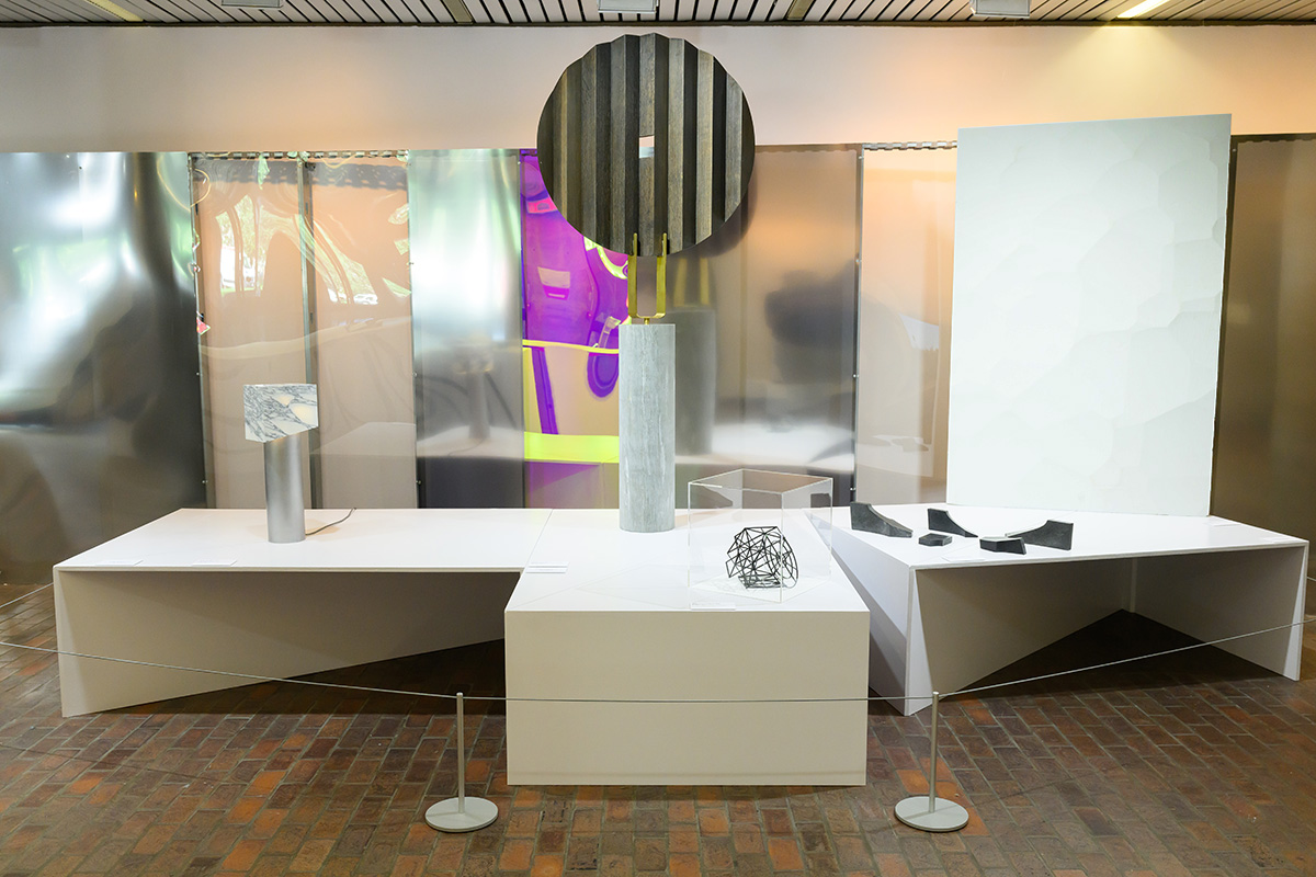 A variety of art objects on display tables in front of a curved reflective plexiglass wall inside Druker Design Gallery.