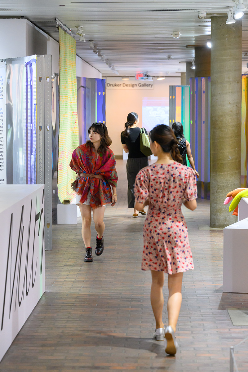 People walking through the exhibit in Druker Design Gallery with curved plexiglass walls on either side.