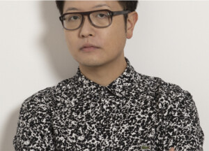 A man with black hair and glasses wearing a black and white paint-splatter pattern button-down shirt is seen chest-up in front of an off-white background