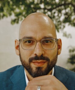Headshot of Andrew Manuel Crespo, who wears glasses and a blue jacket.