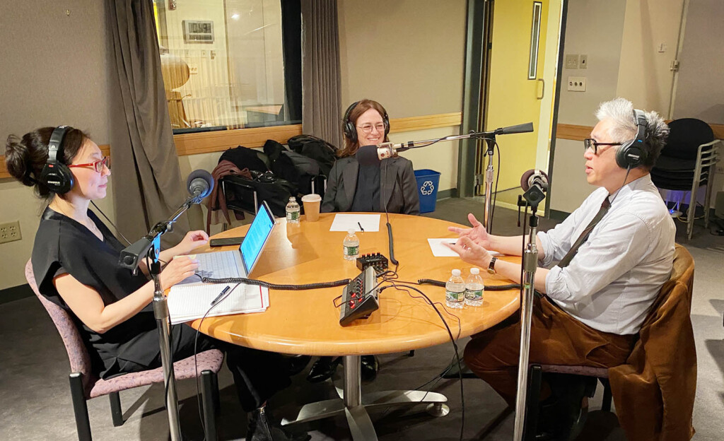 Three people sitting at a round table with headphones on and speaking to microphones