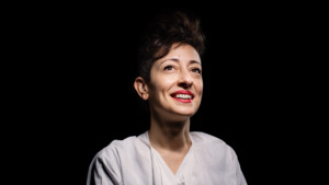 A Lebanese woman with short dark hair and red lipstick wearing a white v-neck blouse against a black background