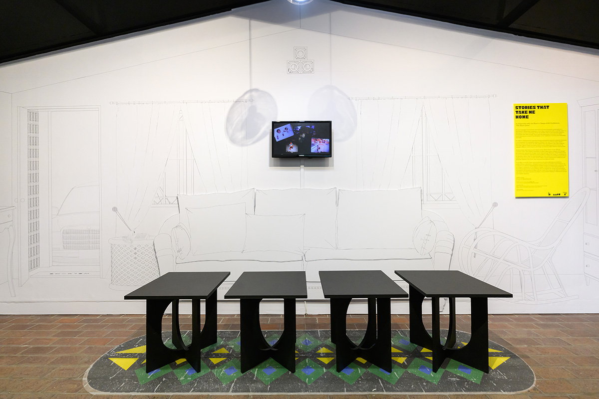 A group of four black stools in front of a line drawing of a couch and rocking chair, with a video screen on the wall showing family photos.