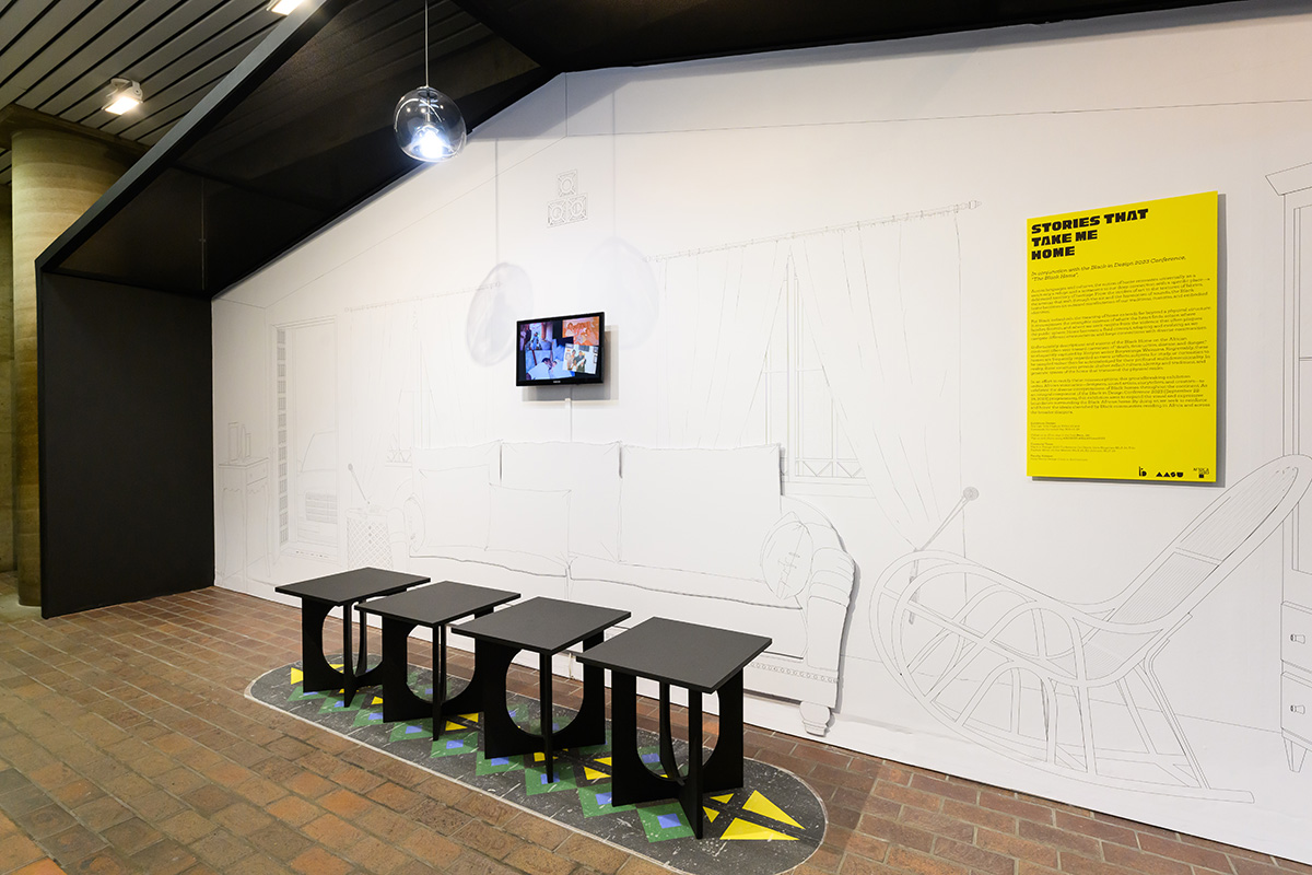 A view of the exhibit in Gund hall showing a group of four stools in front of a line drawing of a domestic living room with video screens on the walls and a roof structure overhead.