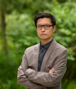 A man with short black hair and black-rimmed glasses stands in front of green foliage. He is wearing a stone-colored blazer over a dark patterned button-down shirt. His arms are folded across his chest and he is seen from waist up. 