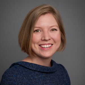 Photograph of a white woman with shortl, light brown hair wearing a navy blue sweater against a grey background.