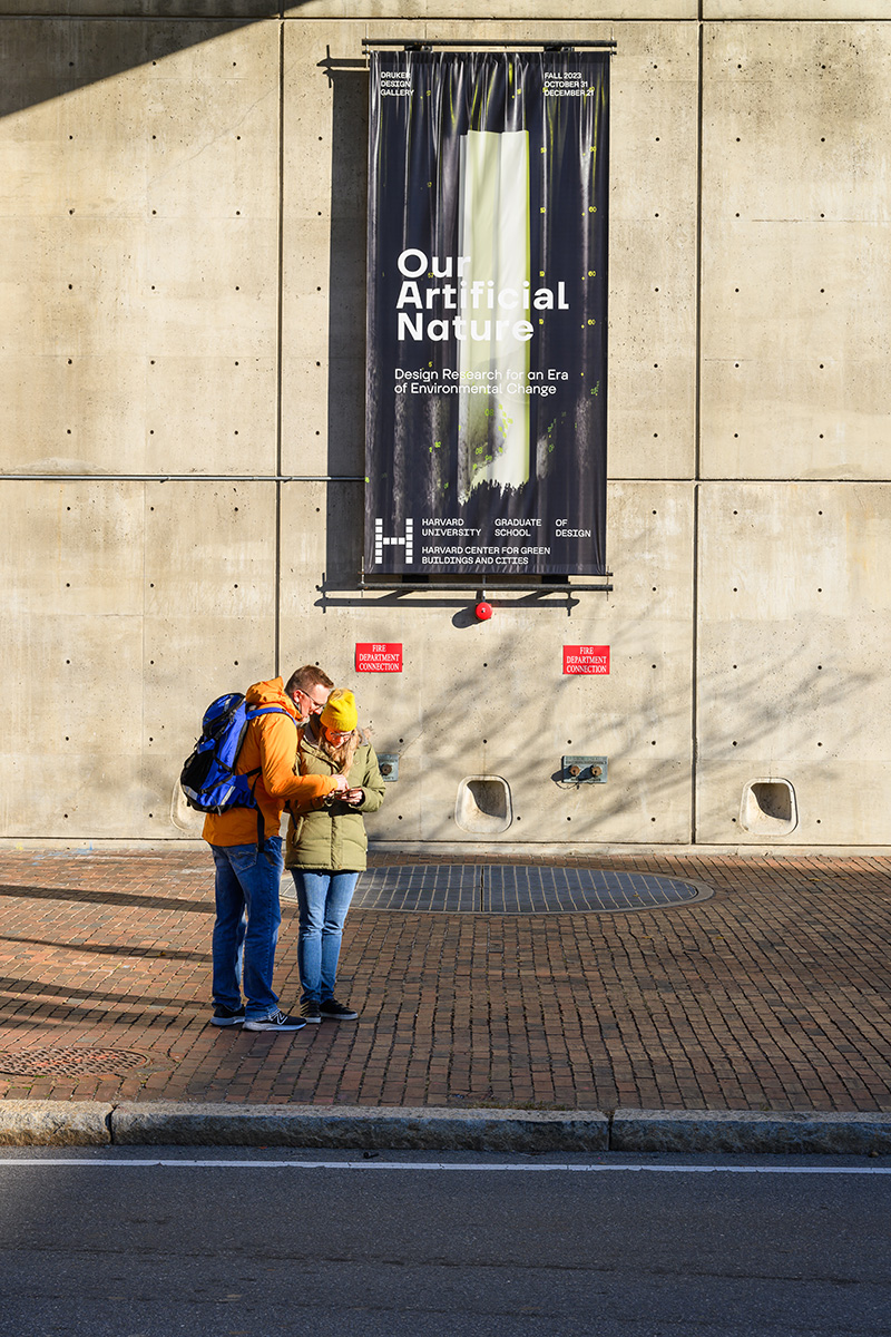 Two people standing outside in front of Gund Hall with the “Our Artificial Nature” banner hanging on the wall behind them.