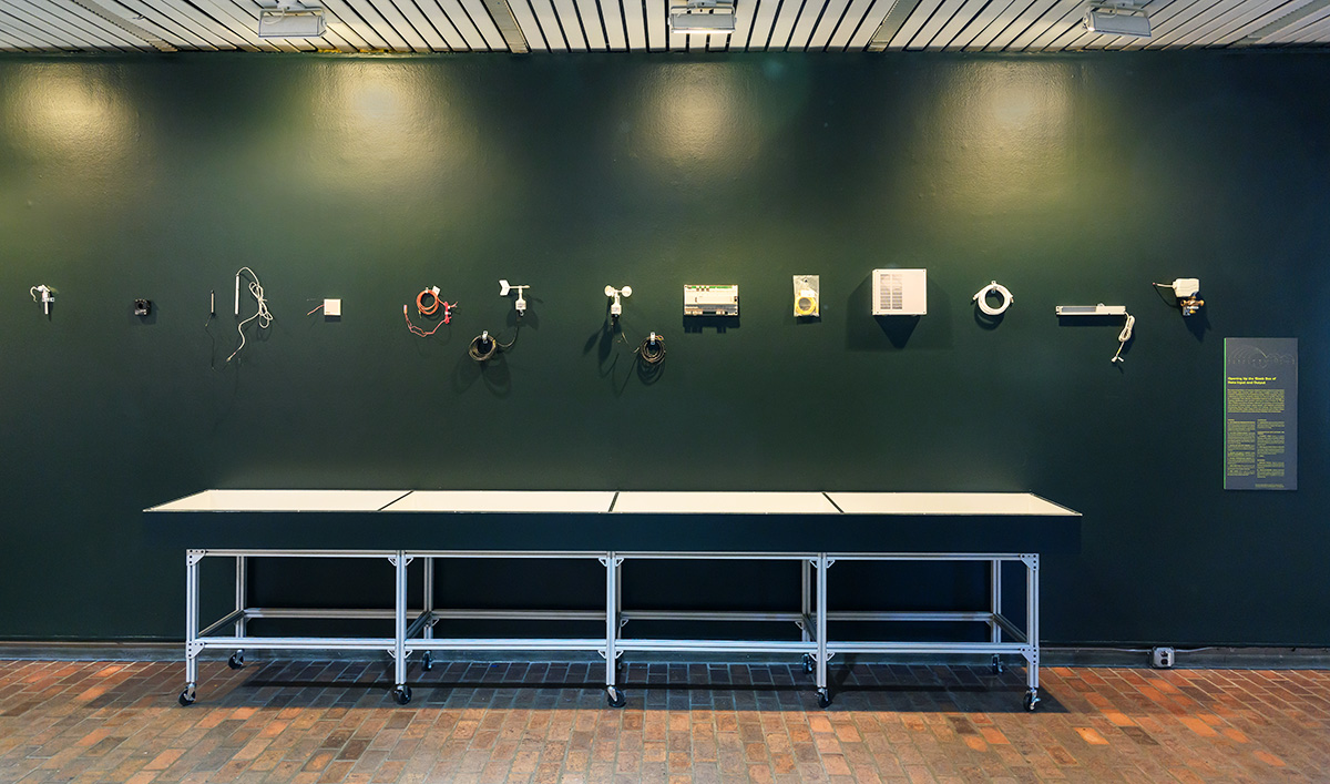 A view inside Druker Design gallery with a variety of electrical and technical hardware displayed on the wall above a long display case.