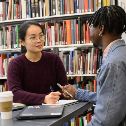 Photo of two students talking at a desk in the library