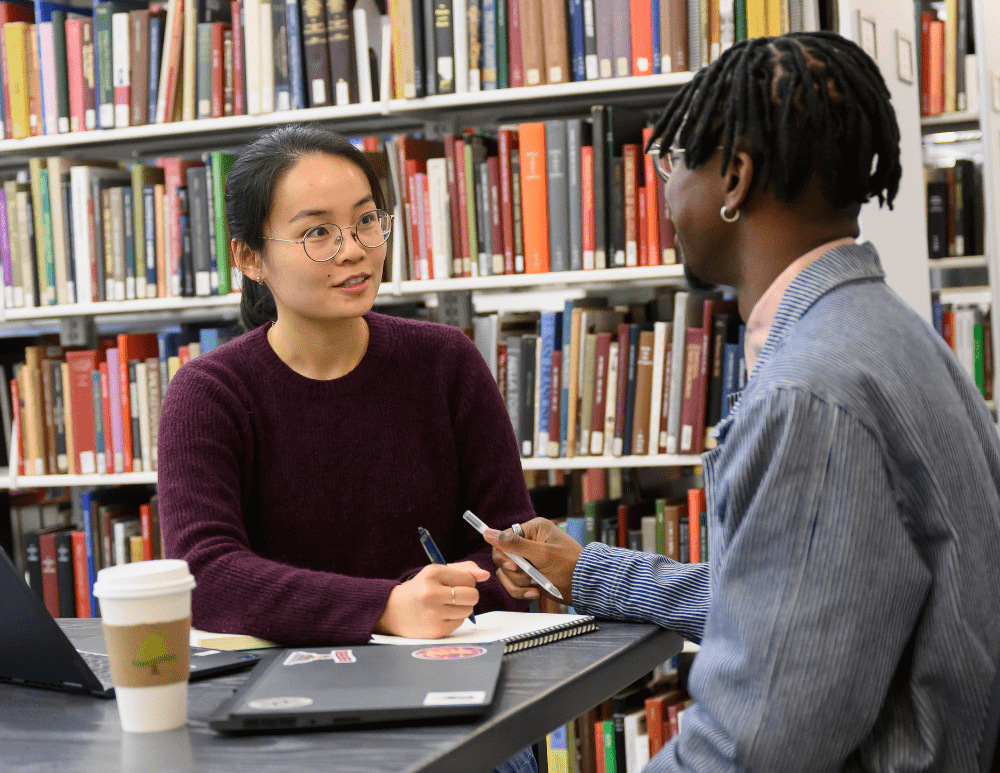 Photo of two students talking at a desk in the library