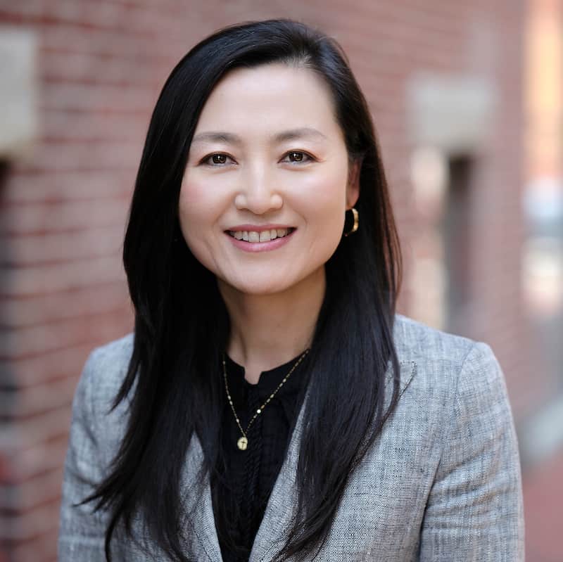 Headshot of Juanne Zhao looking into the camera and smiling while wearing grey suit jacket over a black blouse.