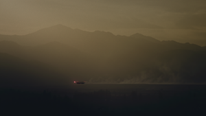 A hazy scene of distant mountains and a building with red lights and smoke coming off it. 