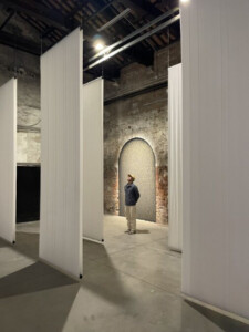 Installation view of "unknown, unknown" at the Venice Architecture Biennale. Long white sheets hang down from the ceiling. A person looks up at the sheets.