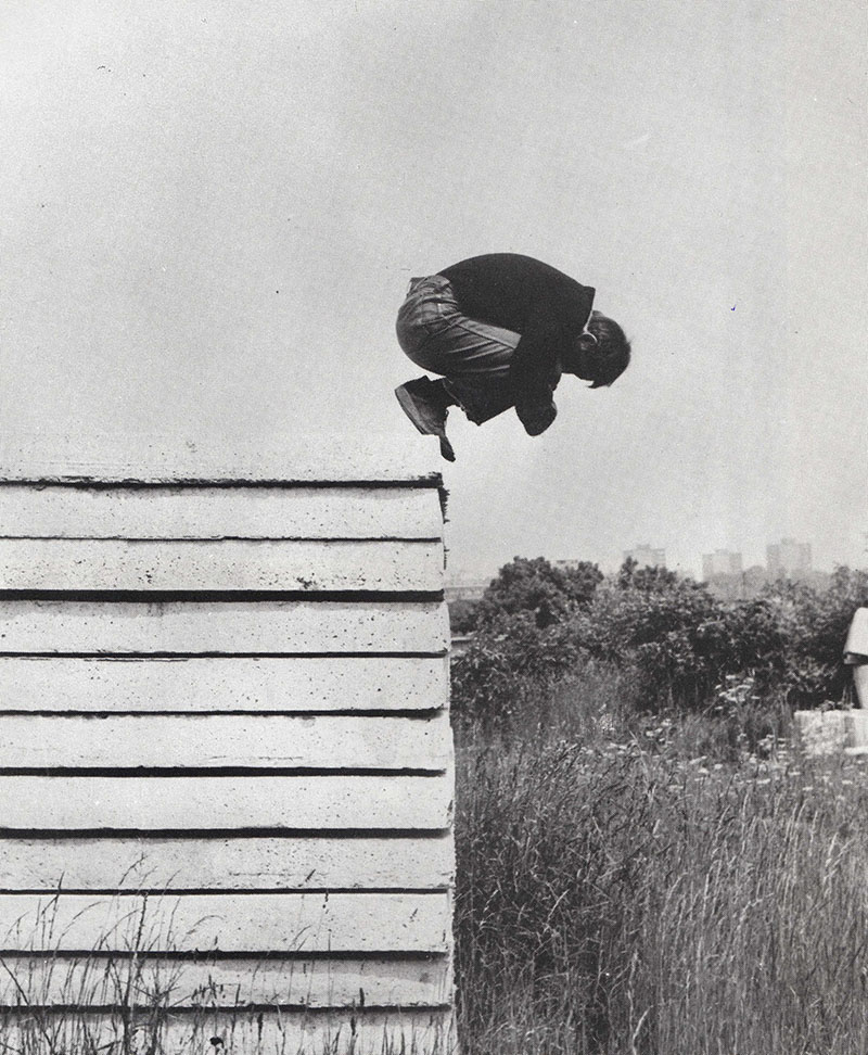 A black-and-white photograph by artist Karel Miler from his Actions series. A man stands on a ledge in a fetal position.