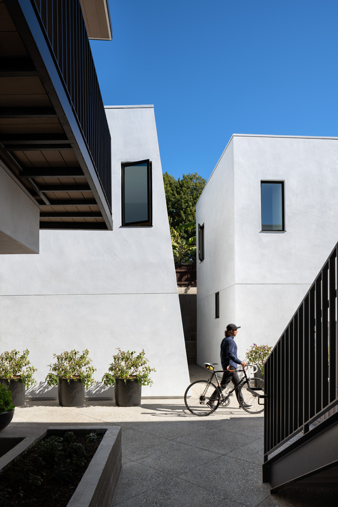 A view of a courtyard surrounding by a cluster of white modernist buildings. A person with a bicycle walks through the courtyard.