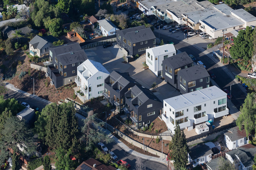 An aerial view of a housing development on a Southern California hillside with a mix of predominantly black and predominantly white two or three story houses clustered around a central street.