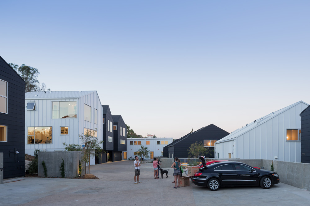 A view of people standing in a shared driveway area with a few cars. They are surrounded by a dense cluster of modernist homes.