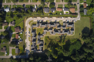 An aerial view of connected townhouses surrounded by green fields and forest. A curving road runs along the townhouse site.