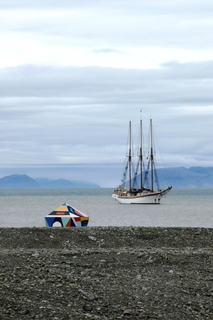 colorful geometric structure on shore of lake with tall ship and mountains in background