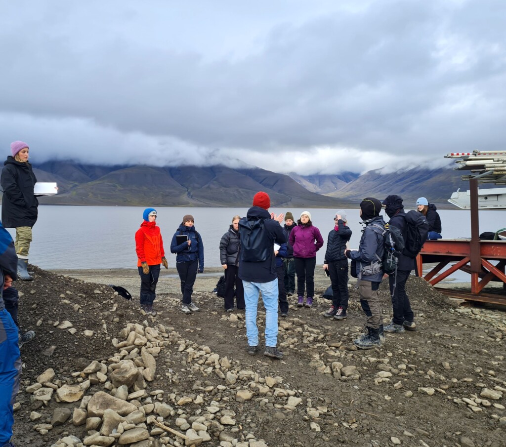 small crowd of people listening to speaker on shore of lake with mountains in background