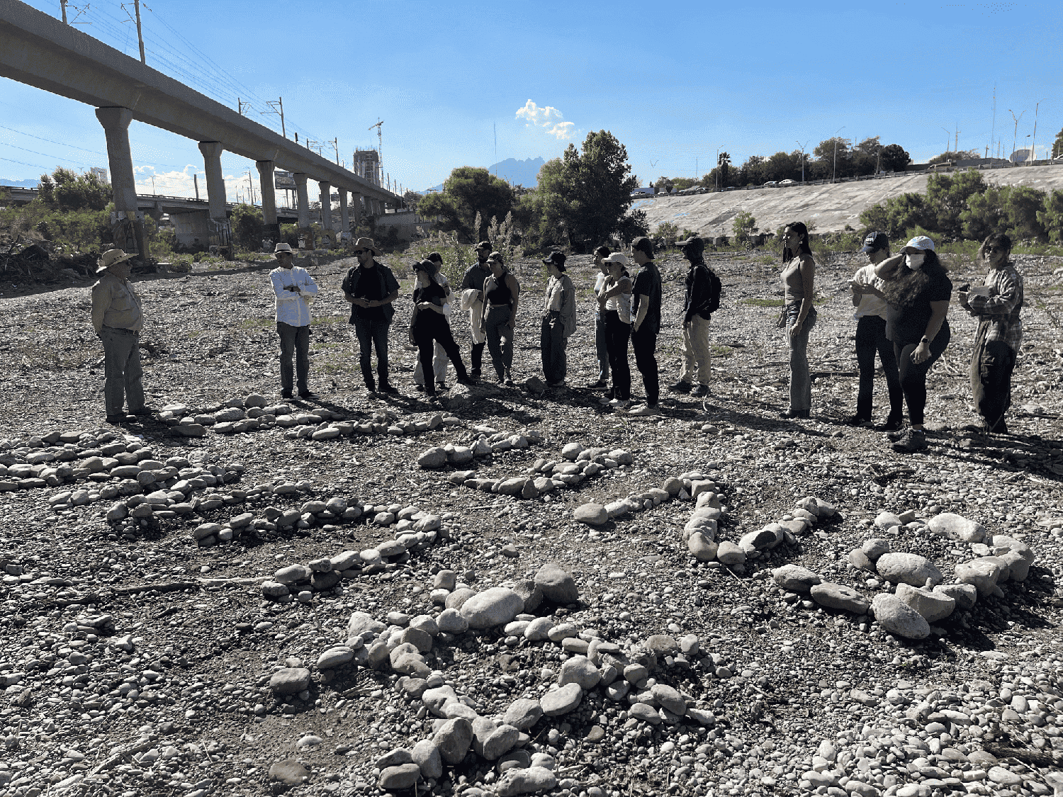 People standing around a dry riverbed. In the background is an elevated highway. In the foreground are stones arranged to form letters.