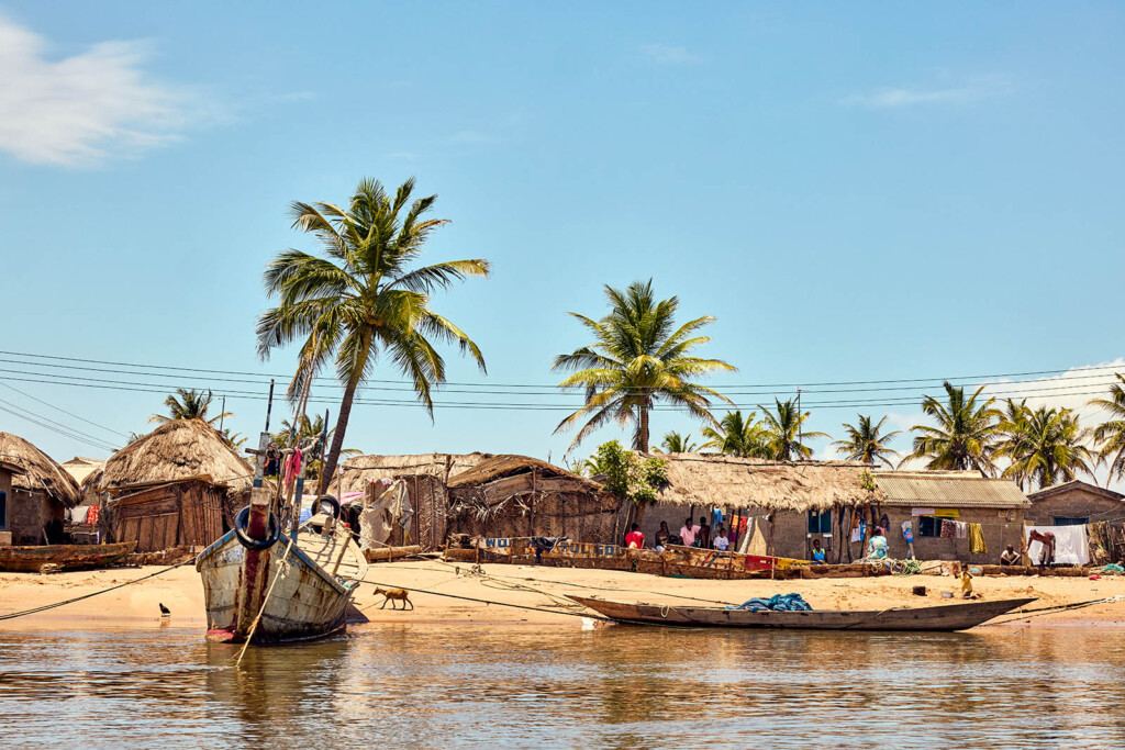 A photo of thatch roofed houses on a bank of a river with two moored boats in the foreground