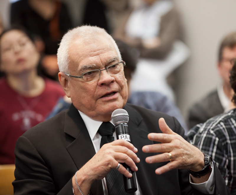 A photograph of architect and scholar George Baird holding a microphone and apparently speaking in a room of other people.