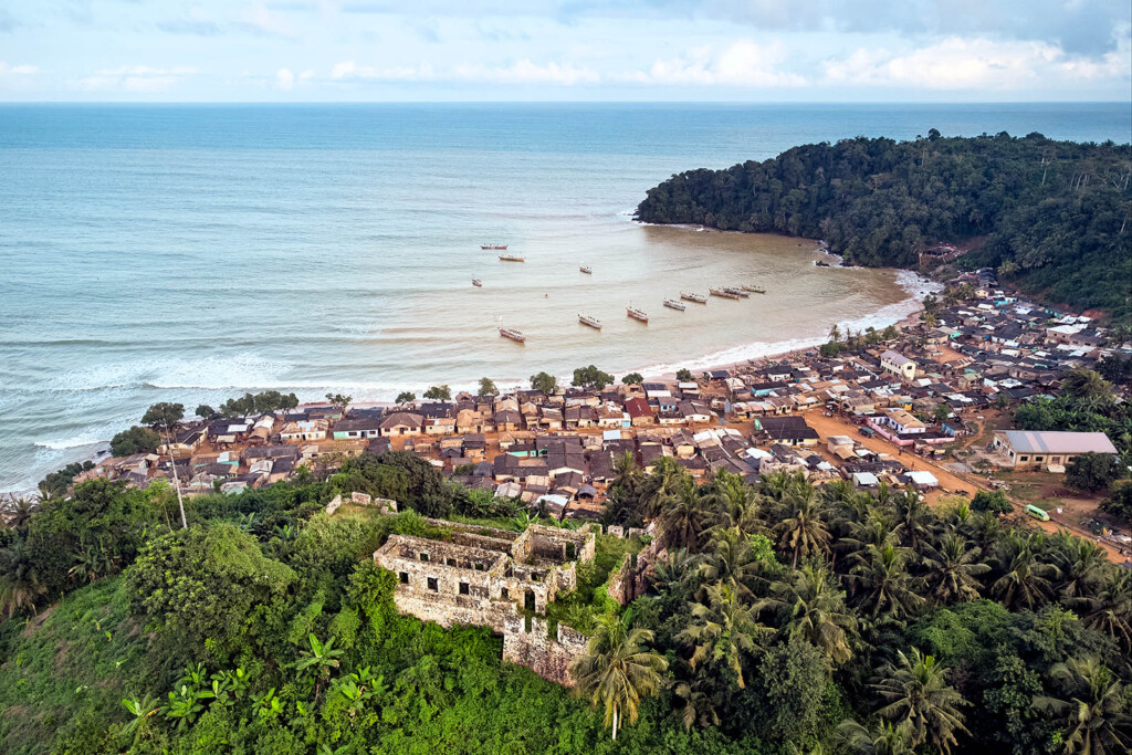 Aerial photo of a densely populated village along the ocean coast with ruins of a building perched on the mountain in the foreground