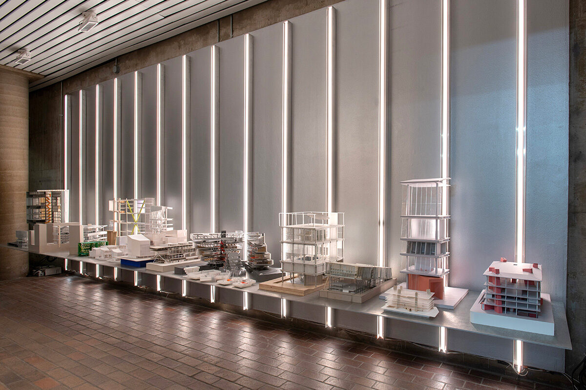 A variety of architectural models on a metal display shelf with vertical lighting behind them.